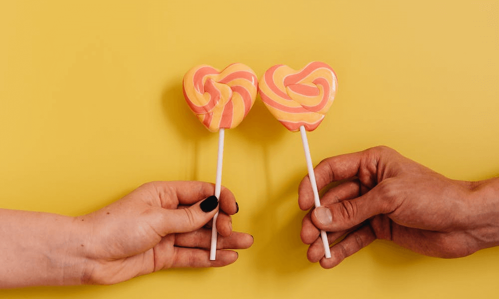 Couple hands holding heart shaped lollipops on yellow background